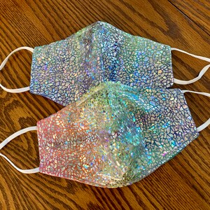 May include: Two face masks with a shimmering, mosaic-like pattern in shades of blue, green, and pink. Each mask has white adjustable ear loops for a comfortable fit. The masks are designed for personal use.