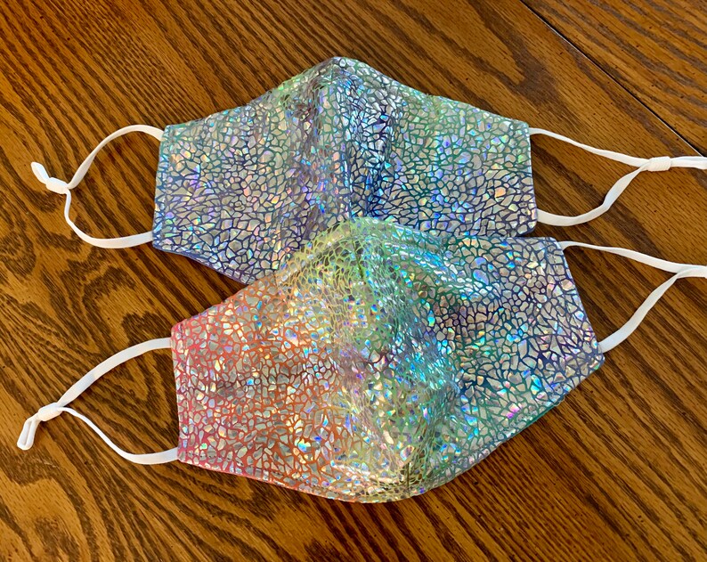 May include: Two face masks with a shimmering, mosaic-like pattern in shades of blue, green, and pink. Each mask has white adjustable ear loops for a comfortable fit. The masks are designed for personal use.