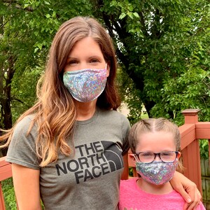 May include: Two face masks with iridescent sequins. One mask is worn by a person wearing a green t-shirt with "The North Face" printed on it. The other mask is worn by a child wearing glasses and a pink shirt.