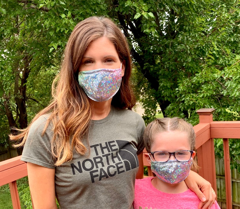 May include: Two face masks with iridescent sequins. One mask is worn by a person wearing a green t-shirt with "The North Face" printed on it. The other mask is worn by a child wearing glasses and a pink shirt.