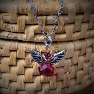 May include: A silver-toned necklace featuring a pendant of an angel with detailed wings and a faceted, deep red crystal body. The chain is delicate, and the pendant is suspended against a woven basket background.