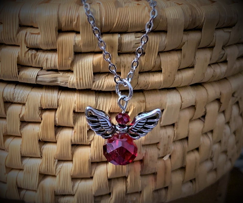 May include: A silver-toned necklace featuring a pendant of an angel with detailed wings and a faceted, deep red crystal body. The chain is delicate, and the pendant is suspended against a woven basket background.