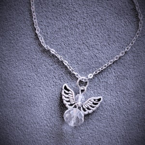 May include: Silver-toned angel pendant necklace. The pendant features an angel with detailed wings and a clear crystal bead body. The necklace chain is delicate and silver, set against a gray background.