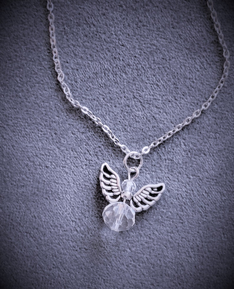 May include: Silver-toned angel pendant necklace. The pendant features an angel with detailed wings and a clear crystal bead body. The necklace chain is delicate and silver, set against a gray background.