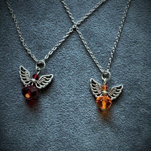 May include: Two silver-toned angel wing pendant necklaces. One features a red faceted bead, the other an orange faceted bead. Both necklaces have delicate silver chains, set against a dark gray background.