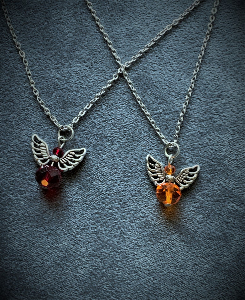 May include: Two silver-toned angel wing pendant necklaces. One features a red faceted bead, the other an orange faceted bead. Both necklaces have delicate silver chains, set against a dark gray background.