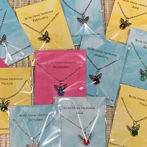 May include: A collection of birthstone necklaces, each displayed on a colorful card. The necklaces feature a silver-toned chain and an angel pendant with a colored gemstone. Cards are labeled with months and associated traits.