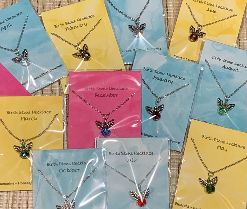 May include: A collection of birthstone necklaces, each displayed on a colorful card. The necklaces feature a silver-toned chain and an angel pendant with a colored gemstone. Cards are labeled with months and associated traits.