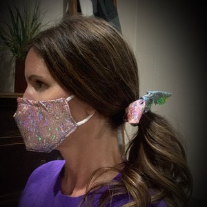 May include: A pink, sparkly face mask with white ear loops is worn. The mask has a textured, cracked pattern. A matching hair tie with a similar sparkly design secures a ponytail. The person is wearing a purple shirt.