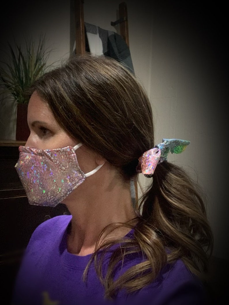 May include: A pink, sparkly face mask with white ear loops is worn. The mask has a textured, cracked pattern. A matching hair tie with a similar sparkly design secures a ponytail. The person is wearing a purple shirt.