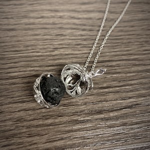 May include: A silver-colored aromatherapy necklace with a decorative filigree pendant, open to reveal a dark, porous lava stone. The necklace chain is delicate and silver. The pendant is open, allowing the scent of essential oils to diffuse.
