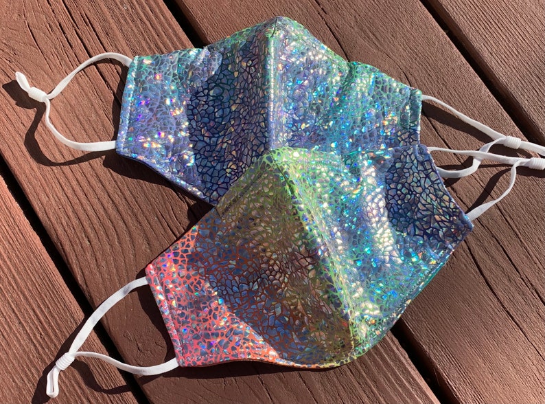 May include: Two face masks with adjustable white ear loops. The masks have a shimmering, iridescent finish with a mosaic-like pattern. One mask transitions from pink to green, while the other is primarily blue.