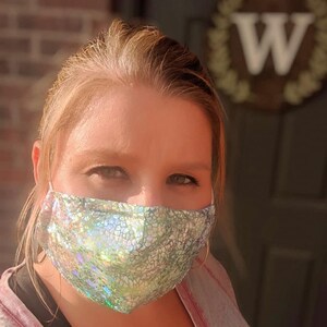 May include: A face mask with a shimmering, iridescent pattern in shades of silver and green. The mask is made of a textured fabric and has white elastic ear loops. The mask covers the nose and mouth.