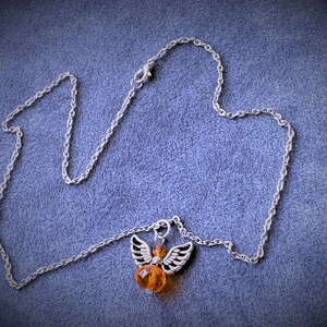 May include: A silver-toned necklace with a pendant featuring an angel design. The angel has silver wings and an orange crystal body. The necklace chain is delicate and rests on a blue-grey surface.