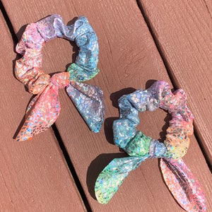 May include: Two iridescent scrunchies with a rainbow color scheme. The scrunchies have a textured, scale-like pattern and are tied with a bow. The colors include pink, orange, yellow, green, blue, and purple.
