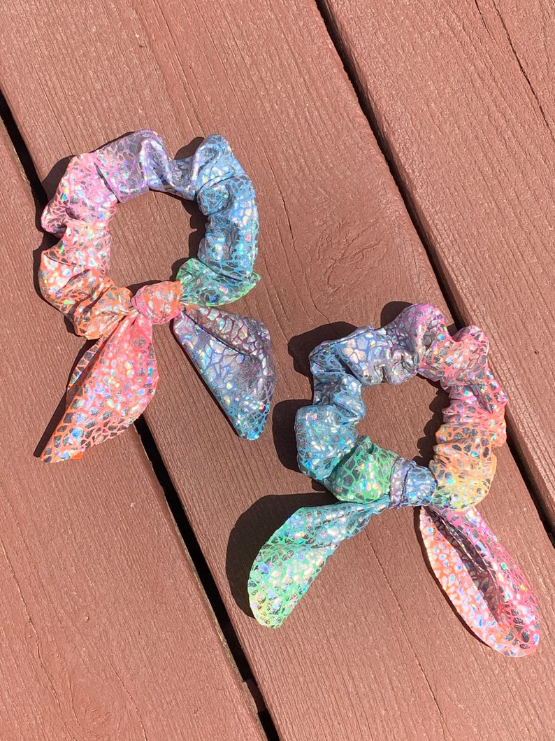 May include: Two iridescent scrunchies with a rainbow color scheme. The scrunchies have a textured, scale-like pattern and are tied with a bow. The colors include pink, orange, yellow, green, blue, and purple.