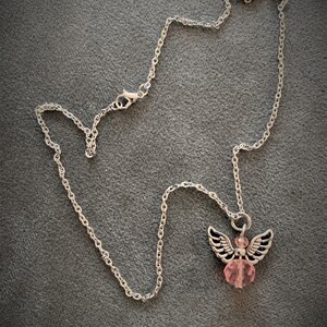 May include: A silver-colored necklace with an angel pendant. The pendant features a pink crystal bead body, silver wings, and a silver halo. The necklace chain is a delicate link style, and the background is a solid gray.