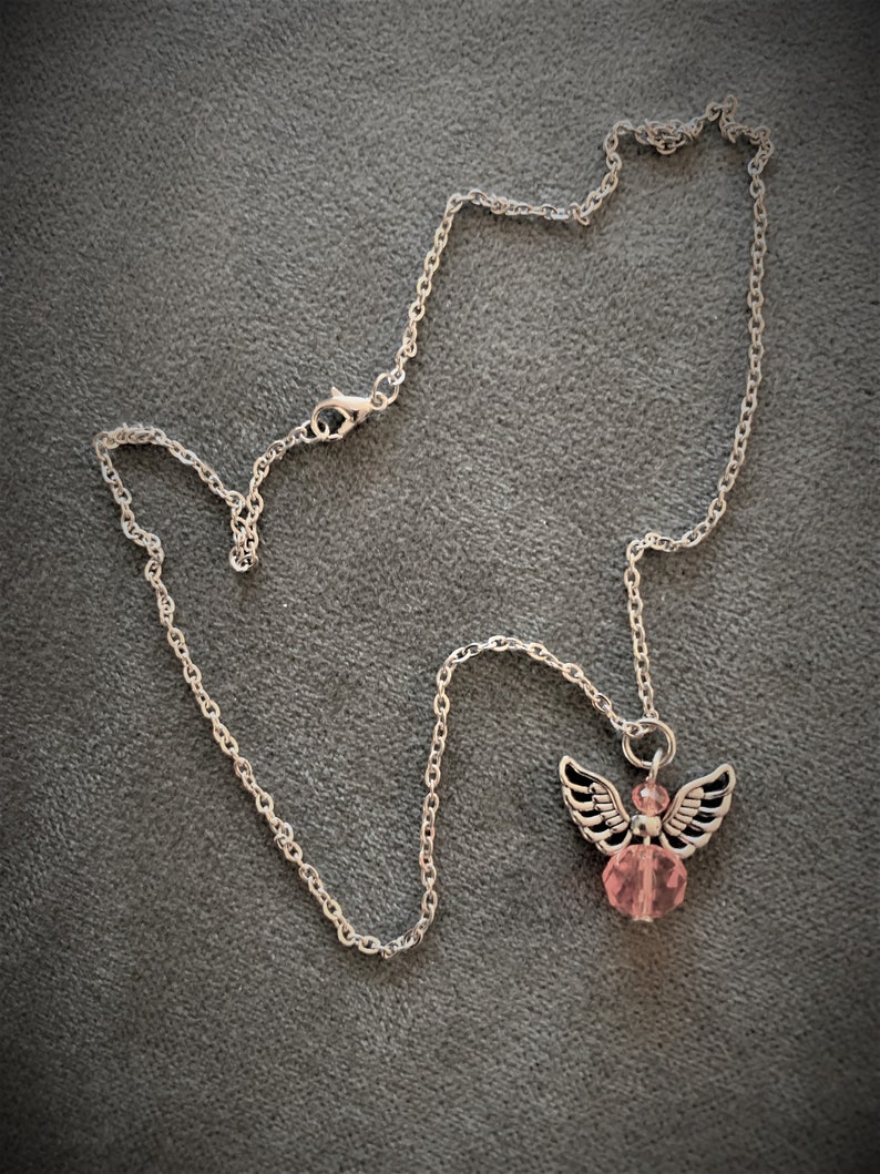 May include: A silver-colored necklace with an angel pendant. The pendant features a pink crystal bead body, silver wings, and a silver halo. The necklace chain is a delicate link style, and the background is a solid gray.