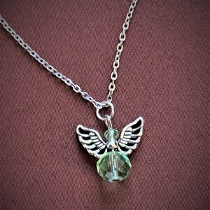 May include: Silver-toned necklace featuring a small angel pendant with detailed wings and a faceted green crystal body. The chain is delicate, and the pendant hangs against a dark red-brown background.