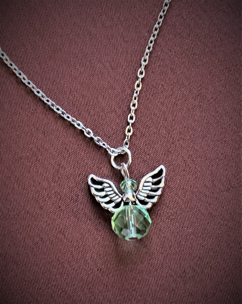 May include: Silver-toned necklace featuring a small angel pendant with detailed wings and a faceted green crystal body. The chain is delicate, and the pendant hangs against a dark red-brown background.