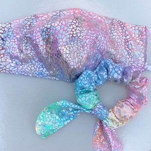 May include: A face mask and matching scrunchie set. Both items feature a shimmering, iridescent pattern in shades of pink, purple, blue, and silver. The mask has white elastic ear loops. The scrunchie is tied in a bow.