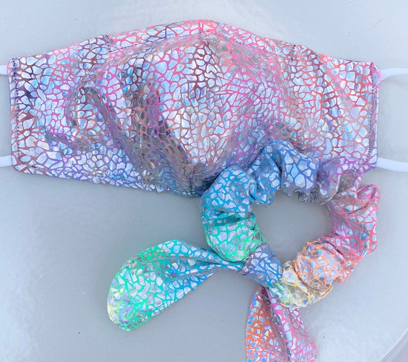 May include: A face mask and matching scrunchie set. Both items feature a shimmering, iridescent pattern in shades of pink, purple, blue, and silver. The mask has white elastic ear loops. The scrunchie is tied in a bow.