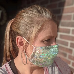 May include: A face mask with a shimmering, iridescent pattern in shades of green and silver. The mask is made of a textured fabric and has white elastic ear loops. The person is wearing large silver hoop earrings.