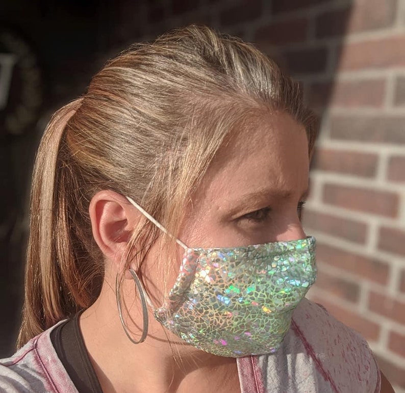 May include: A face mask with a shimmering, iridescent pattern in shades of green and silver. The mask is made of a textured fabric and has white elastic ear loops. The person is wearing large silver hoop earrings.