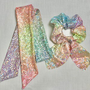 May include: A set of iridescent hair accessories, including a long scarf and a scrunchie with a bow. The items feature a mosaic-like pattern and a color gradient from purple to green, with hints of pink and yellow.