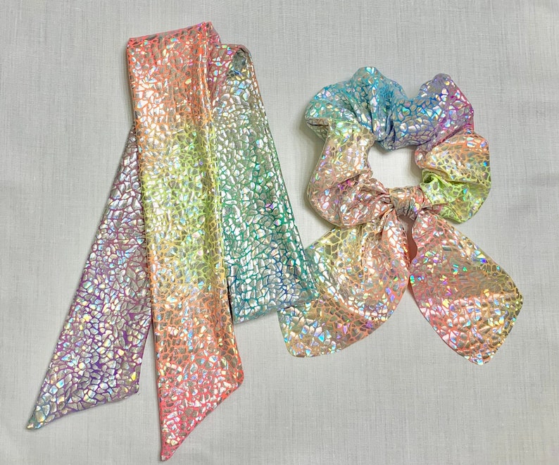 May include: A set of iridescent hair accessories, including a long scarf and a scrunchie with a bow. The items feature a mosaic-like pattern and a color gradient from purple to green, with hints of pink and yellow.