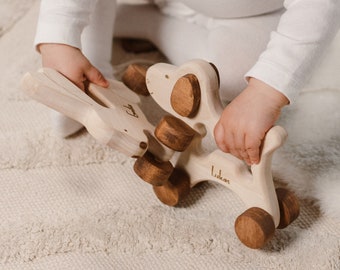May include: A wooden toy dog with brown wheels and details. The toy is engraved with the name "Lukas". The child is wearing a white long-sleeved top with a black trim collar. The toy is made of light-colored wood.