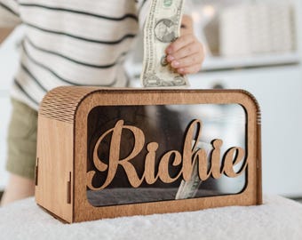 May include: A wooden money box with a clear plastic window and the name "Richie" engraved on the front. A child is putting a dollar bill into the box.