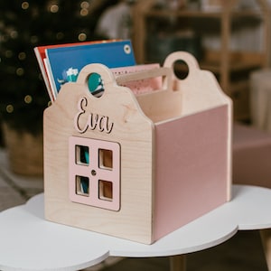 May include: A wooden toy book organizer shaped like a house with a pink interior and a window. The house has a handle on top and the name "Eva" is written on the front.