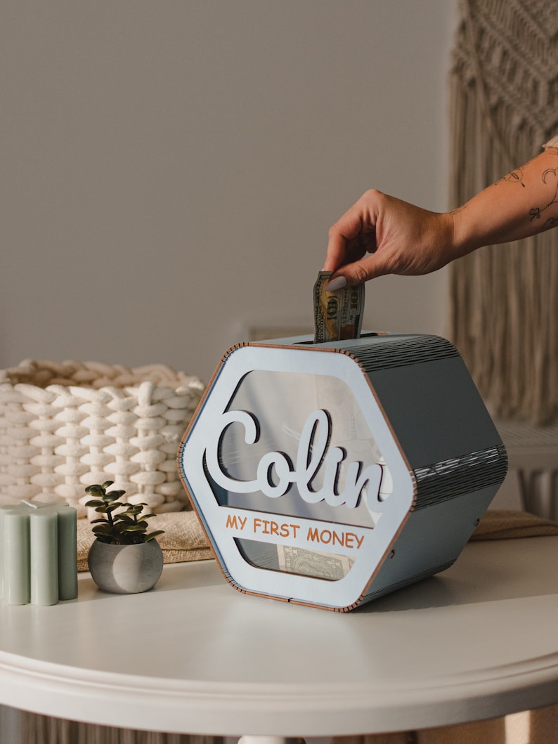 May include: A light blue wooden money box with a clear plastic window. The box has the name "Colin" and the text "My First Money" on the front. A hand is putting a dollar bill into the box.