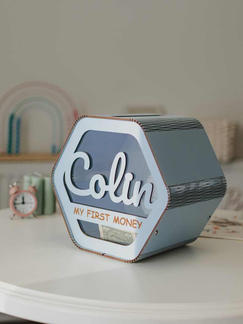 May include: A light blue wooden hexagonal money box with a clear plastic window. The box has the name "Colin" and the text "My First Money" printed on it.
