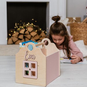 May include: A wooden book storage box shaped like a house with a pink roof and a window. The box has the name "Eva" written on the front.
