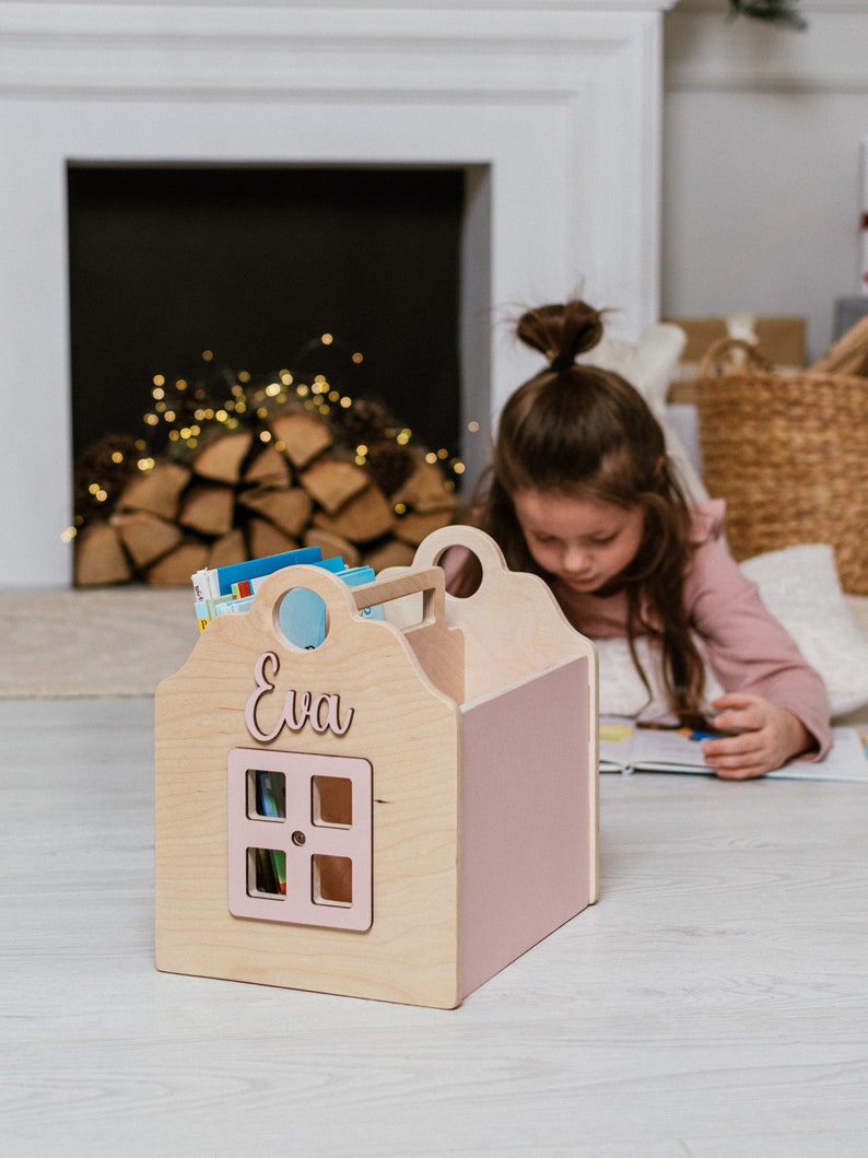 May include: A wooden book storage box shaped like a house with a pink roof and a window. The box has the name "Eva" written on the front.