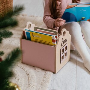 May include: A pink wooden book holder shaped like a house with the name "Eva" on the front. The holder is filled with colorful children's books, including one with the title "Dr. Seuss".