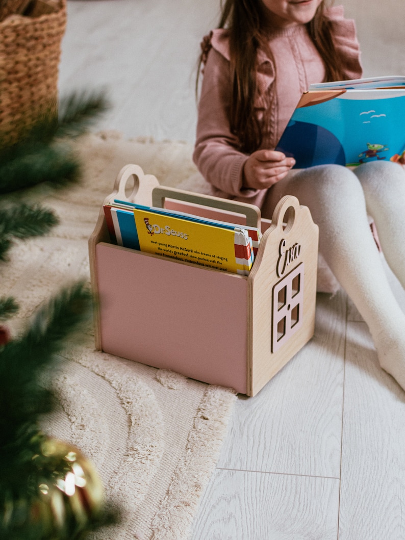 May include: A pink wooden book holder shaped like a house with the name "Eva" on the front. The holder is filled with colorful children's books, including one with the title "Dr. Seuss".