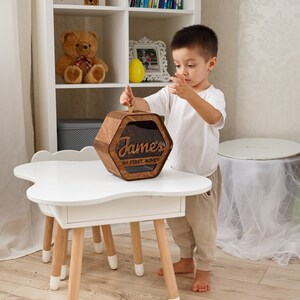 May include: A wooden piggy bank with a hexagonal shape and a clear plastic window. The piggy bank has the name "James" and the text "My First Money" engraved on it. A young child is putting a coin into the piggy bank.