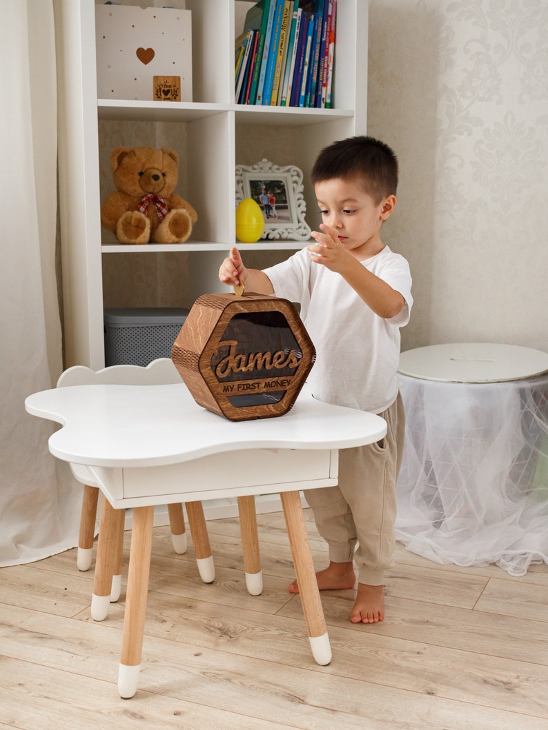 May include: A wooden piggy bank with a hexagonal shape and a clear plastic window. The piggy bank has the name "James" and the text "My First Money" engraved on it. A young child is putting a coin into the piggy bank.
