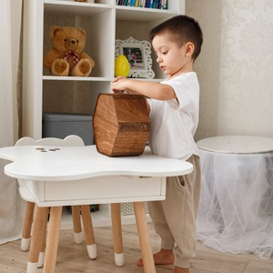 May include: A young child in a white shirt and beige pants is putting money into a wooden hexagonal piggy bank. The piggy bank is sitting on a white table with a drawer. The table has four wooden legs with white tips.