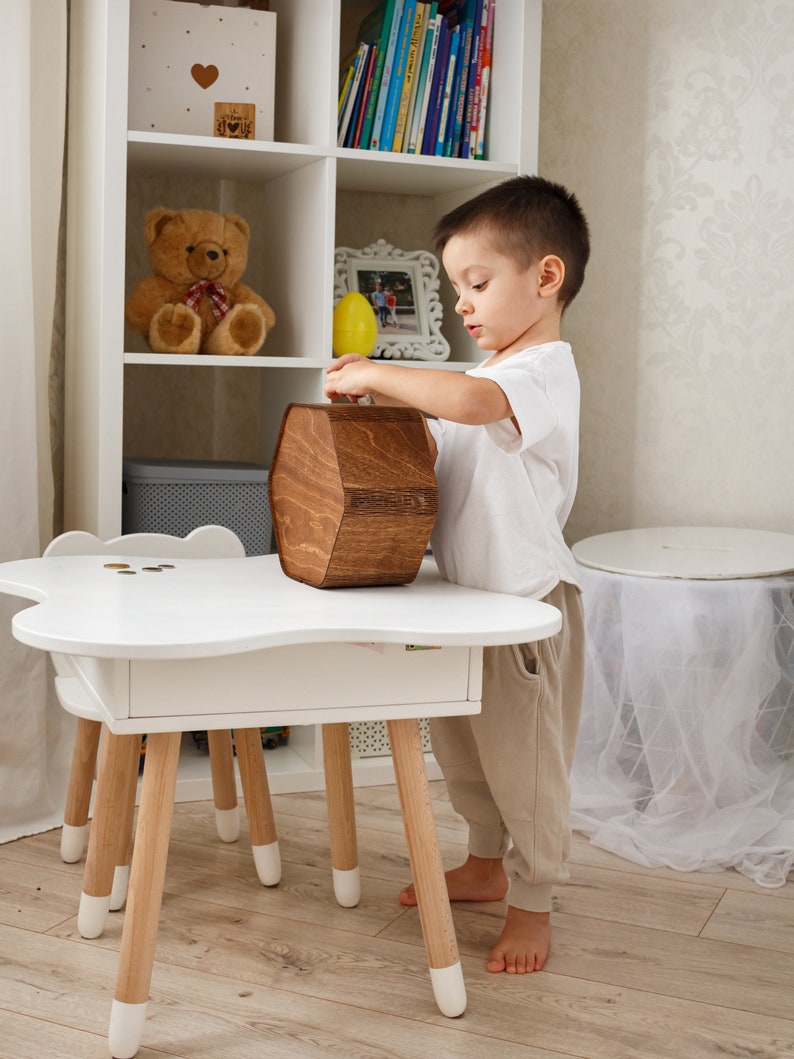 May include: A young child in a white shirt and beige pants is putting money into a wooden hexagonal piggy bank. The piggy bank is sitting on a white table with a drawer. The table has four wooden legs with white tips.