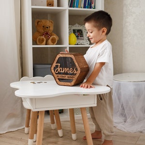 May include: A wooden hexagonal piggy bank with a slot on top. The piggy bank is personalized with the name "James" and the text "My First Money". A young child is holding the piggy bank in front of a white table with wooden legs.