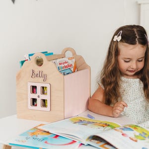 Könnte beinhalten: Ein Bücherregal aus Holz in Hausform mit dem Namen "Betty" auf der Vorderseite. Das Bücherregal hat eine rosa Tür und enthält mehrere Bücher. Ein junges Mädchen sitzt in der Nähe und betrachtet ein offenes Buch auf einem weißen Tisch. Im Hintergrund hängt eine Wimpelkette.
