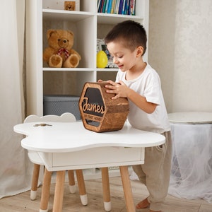 May include: A wooden piggy bank with the name "James" and the text "My First Money" engraved on it. A young boy is holding the piggy bank in a white room with a white bookshelf in the background.