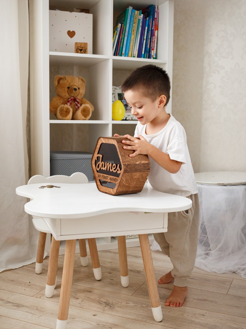 May include: A wooden piggy bank with the name "James" and the text "My First Money" engraved on it. A young boy is holding the piggy bank in a white room with a white bookshelf in the background.