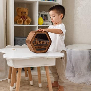 May include: A wooden hexagonal piggy bank with a clear plastic window. The piggy bank is engraved with the name "James" and the words "My First Money". A young child is holding the piggy bank.