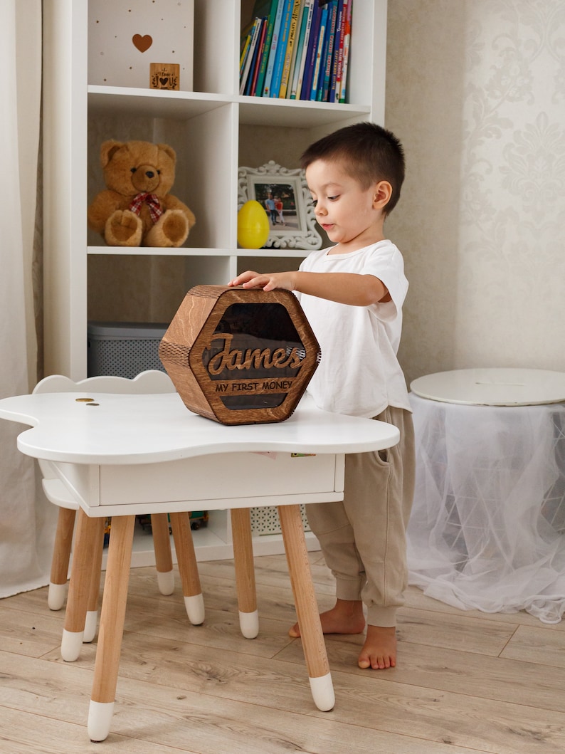 May include: A wooden hexagonal piggy bank with a clear plastic window. The piggy bank is engraved with the name "James" and the words "My First Money". A young child is holding the piggy bank.