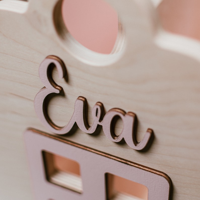 May include: A wooden house-shaped box with the name "Eva" written in rose gold lettering. The box has a small window and a series of smaller windows below the name.